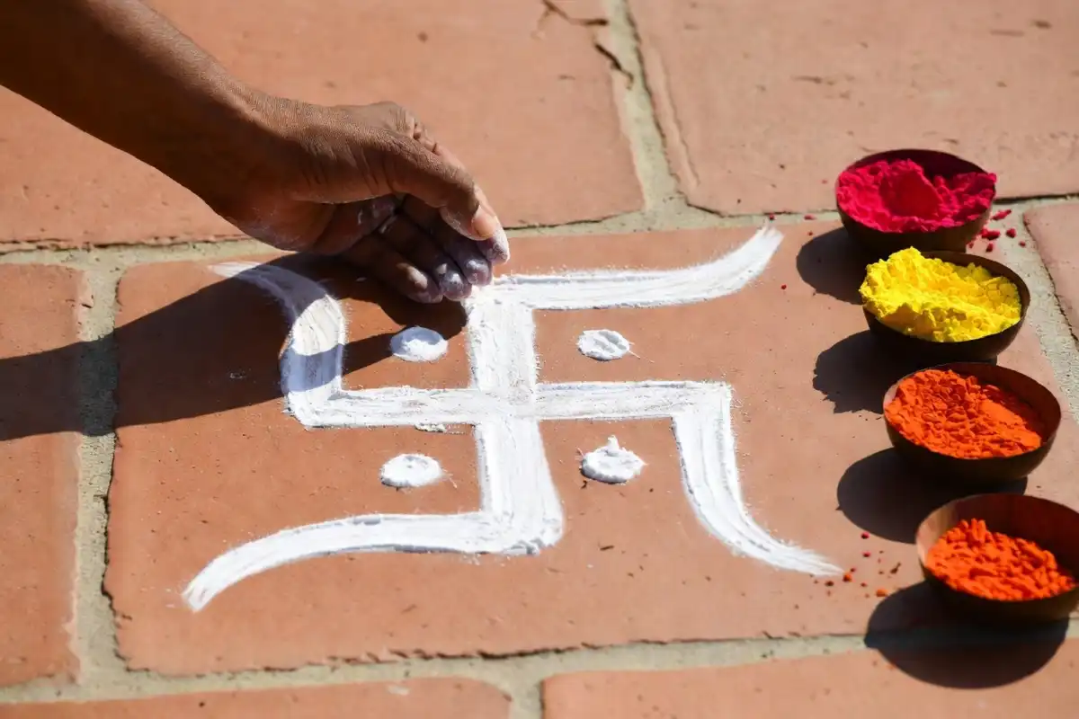 A hand drawing a small swastik rangoli outline in white powder on a floor with colored powder bowls nearby