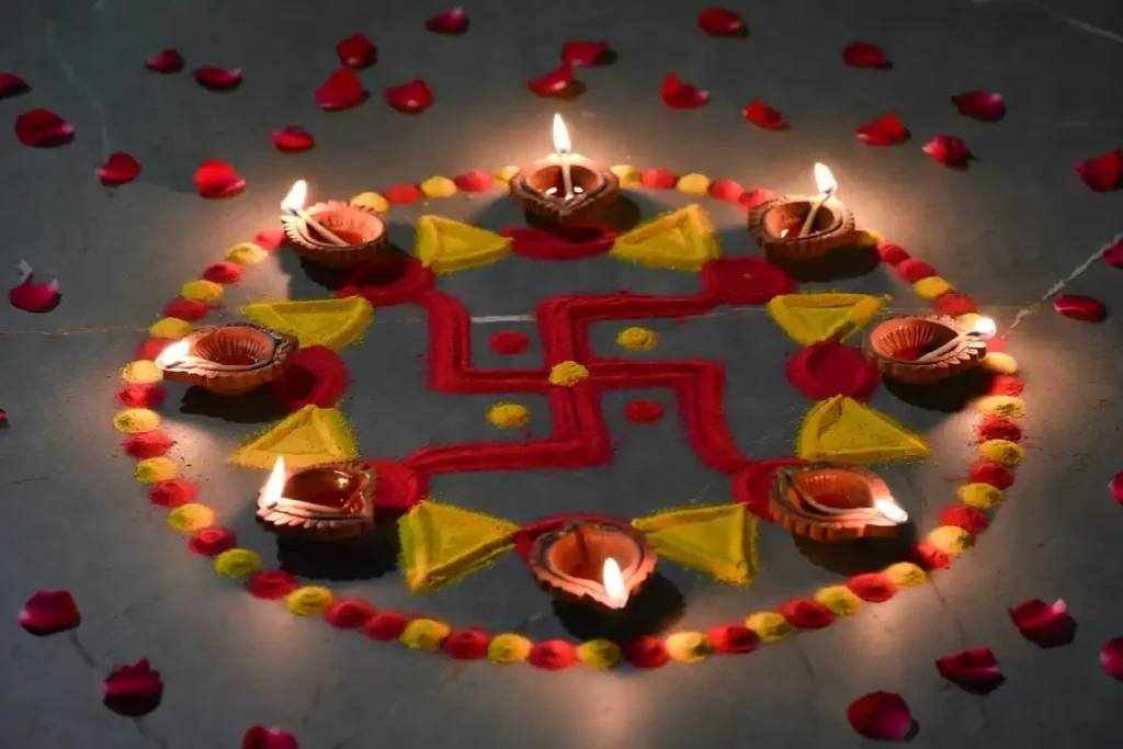 A finished small swastik rangoli in red and yellow surrounded by lit diyas and rose petals on a stone floor