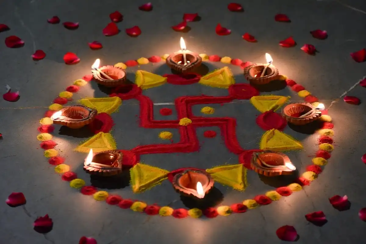 A finished small swastik rangoli in red and yellow surrounded by lit diyas and rose petals on a stone floor